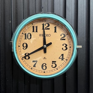 May include: A round Seiko wall clock with a light blue bezel. The clock face is tan with black numerals and hands. The clock reads 2:00. The word "SEIKO" is printed on the clock face. The clock is mounted on a black surface.