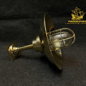 May include: A brass nautical-style wall sconce with a protective wire cage around the glass bulb. The sconce has a flared shade and a square base. The text "VERDIGRISFORGE" is visible.