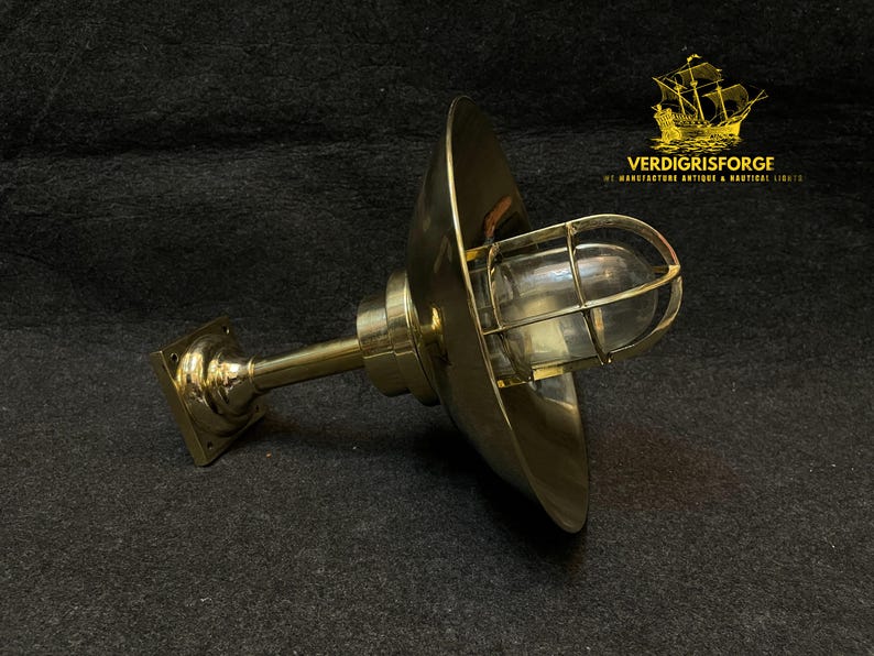 May include: A brass nautical-style wall sconce with a protective wire cage around the glass bulb. The sconce has a flared shade and a square base. The text "VERDIGRISFORGE" is visible.