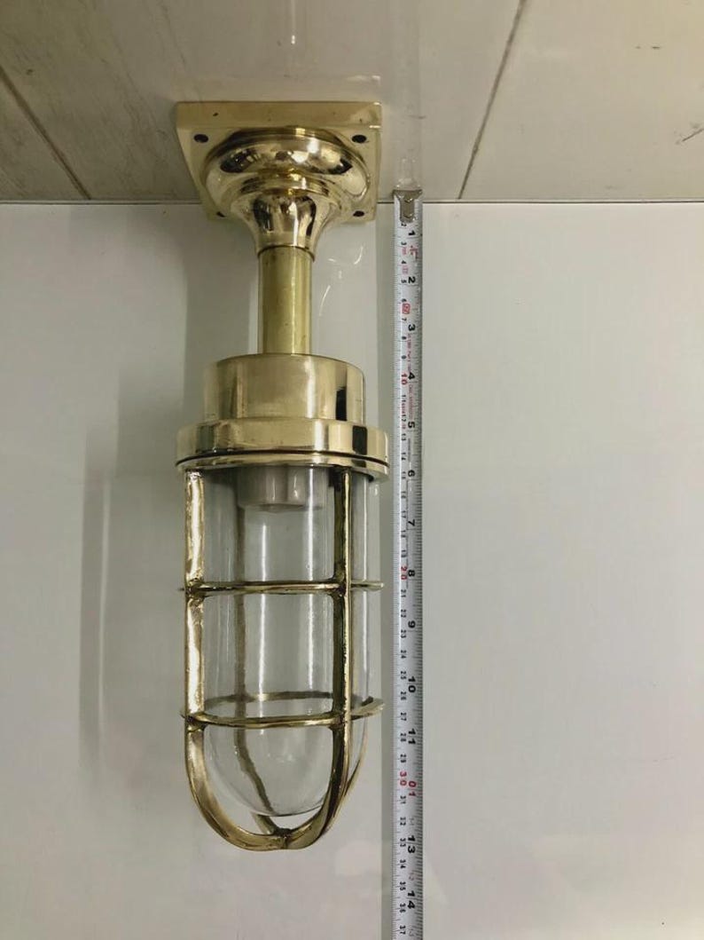 May include: A brass-colored, nautical-style wall-mounted light fixture. The fixture has a clear glass enclosure protected by a metal cage. The light is approximately 14 inches (35.5 cm) tall, with a square mounting plate and a cylindrical body.
