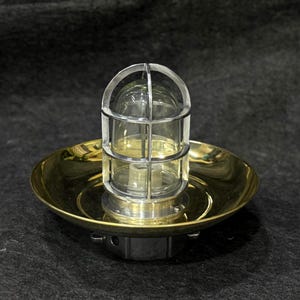 Puede incluir: Una lámpara de latón y vidrio de estilo vintage. La lámpara presenta una carcasa de vidrio transparente encerrada en una jaula de metal, montada sobre una base circular de latón. La luz está diseñada para montarse en una pared o techo.