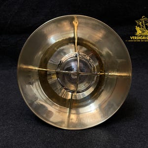 May include: A brass nautical-style light fixture with a round, flared shade. The fixture has a clear glass lens protected by a metal grid. The light is a warm gold color, and the overall design is reminiscent of vintage maritime lighting.