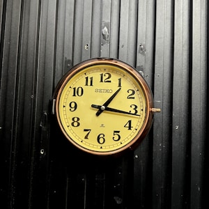 May include: A round Seiko clock with a cream face and black numerals. The clock has a bronze bezel and black hands. It is mounted on a black, vertically-lined surface.