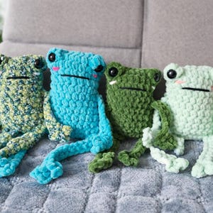 Könnte beinhalten: Vier handgefertigte Häkelfrosch-Plüschtiere in verschiedenen Farben: grün mit gelben Sprenkeln, hellblau, dunkelgrün und hellgrün. Jeder Frosch hat große schwarze Augen, einen schwarzen Mund und rosa Wangen. Sie sitzen auf einer strukturierten grauen Oberfläche.