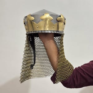 May include: A medieval helmet with a silver-coloured, conical top and a gold-coloured crown-like band. The helmet has a chainmail neck guard. It is held by a person's arm. Likely for historical reenactment or costume use.
