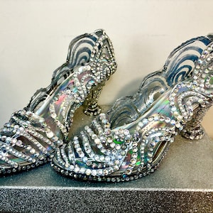 Op de afbeelding: Een paar zilveren pumps met hoge hakken en ingewikkelde strass-versieringen. De open-teen hakken hebben een wervelend ontwerp aan de zijkanten en staan op een glinsterend oppervlak, wat een glamoureuze, formele stijl laat zien.