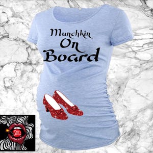 May include: Light blue maternity t-shirt with the text "Munchkin On Board" in black script. The shirt features a pair of red glittery shoes. A small logo is in the lower left corner.