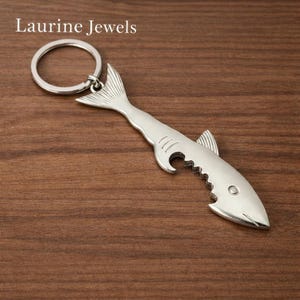 May include: A silver-colored shark-shaped bottle opener with a key ring. The bottle opener has a fin, a tail, and a mouth with jagged teeth. The text "Laurine Jewels" is in the upper left corner.