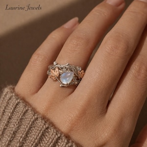 Puede incluir: Anillo plateado con una piedra preciosa azul claro en forma de pera. El anillo presenta un diseño inspirado en la naturaleza con ramas y hojas de color cobre. La marca "Laurine Jewels" es visible en la esquina superior izquierda.