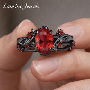 May include: A black ring with a large oval red gemstone as the centerpiece. The ring features a floral design with smaller red gemstones and the text "Laurine Jewels" in the upper left corner.