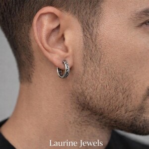 Puede incluir: Un pendiente de aro plateado con un patrón ornamental y ondulado. El pendiente es de diseño circular. El texto "Laurine Jewels" está en la parte inferior de la imagen.