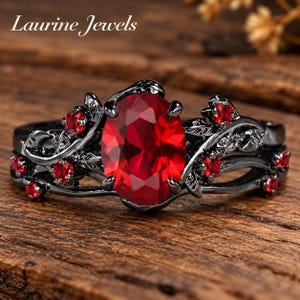 May include: A black metal ring featuring a large oval-cut red centre stone and smaller round red stones set along a band designed with leaf and vine motifs. The text Laurine Jewels is visible in the upper left corner of the image.