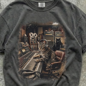 Puede incluir: Una camiseta gris carbón con un estampado gráfico de un gato atigrado con auriculares operando una gran consola de mezcla de audio en un estudio de grabación, con un gato negro sentado sobre un amplificador Marshall al fondo.