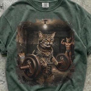 Könnte beinhalten: Ein dunkelgrünes T-Shirt mit einem Grafikdruck einer getigerten Katze, die eine Goldkette trägt und eine schwere Langhantel in einem Fitnessstudio hebt. Eine zweite muskulöse Katze steht im Hintergrund unter einer einzelnen Glühbirne.