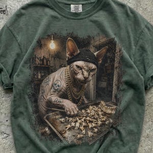 May include: A sage green t-shirt featuring a graphic print of a hairless cat wearing a black beanie and gold chain necklaces. The cat has tattoos of a skull and money bags, and is using a metal tool to sort through a tray of human teeth.