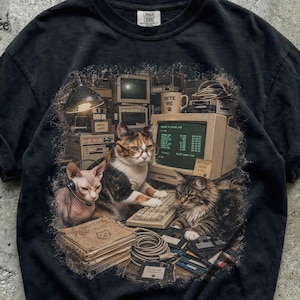 Könnte beinhalten: Ein schwarzes T-Shirt mit einem Grafikdruck von drei Katzen in einem Büro. Eine Katze trägt eine Brille am Computer, eine andere trägt ein Headset und eine dritte sitzt daneben. Der Druck zeigt eine Tasse mit "BYTE ME" und ein Notizbuch mit "SYSTEM CAT-ALOG".