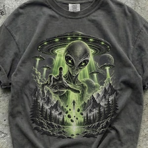 May include: A charcoal grey t-shirt featuring a graphic print of a large alien head centred beneath a glowing UFO. The design includes a mountainous landscape with pine trees, floating rocks, and a small human figure illuminated by a green light beam.