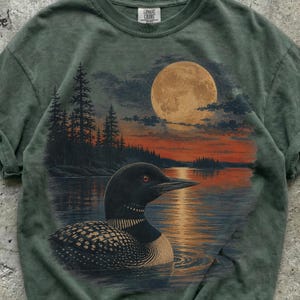 Könnte beinhalten: Ein salbeigrünes T-Shirt mit einem Grafikdruck eines Eistauchers, der bei Sonnenuntergang auf einem See schwimmt. Die Szene zeigt einen großen Vollmond, dunkle Silhouetten von Kiefern und die Spiegelung des Mondes auf dem Wasser. Rundhalsausschnitt.