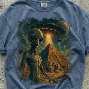 Könnte beinhalten: Ein blaues T-Shirt mit einem Grafikdruck eines grauen Außerirdischen vor einer großen Steinpyramide. Über der Pyramide strahlt eine fliegende Untertasse einen Lichtstrahl aus, während weitere Untertassen und ein Planet am dunklen Himmel zu sehen sind.