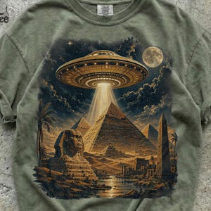 Op de afbeelding: Een olijfgroen t-shirt met een grafische print van een grote UFO die boven de piramides van Gizeh, de Sfinx en een obelisk zweeft onder een sterrenhemel met een volle maan. Het t-shirt heeft een ronde hals en een Comfort Colors label.