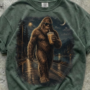 May include: A sage green t-shirt featuring a graphic print of a brown, furry Bigfoot creature wearing sunglasses and holding a plastic cup with a straw. The scene depicts the creature walking along a road near a lake, forest, and crescent moon.