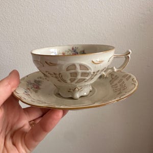 May include: Cream-coloured teacup and saucer set with floral and gold accents. The teacup features a decorative design and a small handle. The saucer has a matching design. A classic tea set.