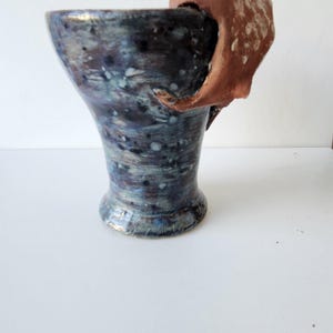 May include: A handmade ceramic vase with a flared top and a unique handle. The vase features a dark blue and purple glaze with white speckles. The handle is a contrasting reddish-brown color.