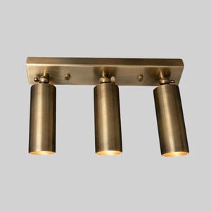 May include: A brass-toned, three-light fixture with cylindrical spotlights. The lights are mounted on a rectangular base. Each spotlight is angled downward, emitting a warm glow. The fixture is designed for ceiling or wall mounting.