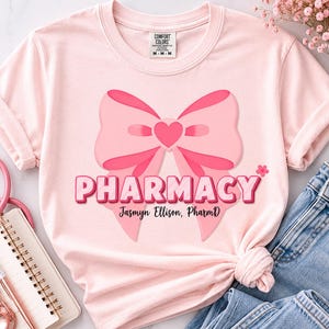 May include: A light pink t-shirt featuring a large pink bow graphic with a central heart. The text PHARMACY is printed in bold pink letters below the bow, followed by the name Jasmyn Ellison, PharmD in a black script font. The shirt has a crew neck.