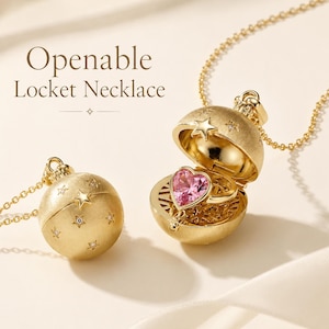 Könnte beinhalten: Eine goldfarbene, kugelförmige Medaillon-Halskette mit matter Oberfläche und sternförmigen Gravuren. Ein Medaillon ist geschlossen, das andere ist geöffnet und zeigt einen rosa herzförmigen Ring. Der Text "Openable Locket Necklace" ist zu sehen.