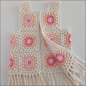 May include: A cream-coloured crochet vest with a floral pattern in pink and peach. The vest has a square neckline and armholes, with a fringe detail along the bottom edge. The open front design adds a touch of bohemian flair.