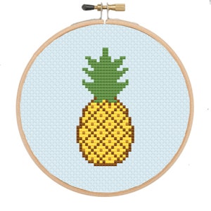 Puede incluir: Una piña amarilla y marrón con la parte superior verde, bordada en una tela azul claro en un aro de bordado de madera redondo.