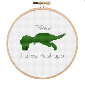 May include: A green T-Rex dinosaur doing pushups with the text "T-Rex Hates Pushups" on a white background in a wooden embroidery hoop.