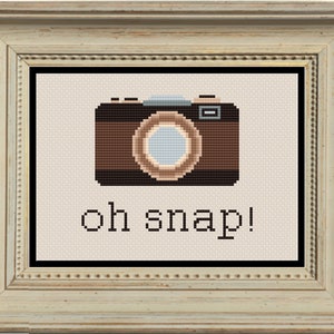 Könnte beinhalten: Ein gerahmtes Kreuzstich-Design mit einer braunen Vintage-Kamera und dem Text "oh snap!" in Schwarz.