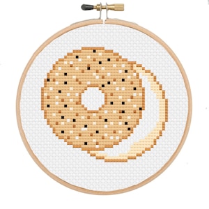 May include: A cross-stitch embroidery hoop featuring a bagel design. The bagel is tan with black and brown speckles, and a cream-colored smear. The embroidery is set in a wooden hoop against a white background.