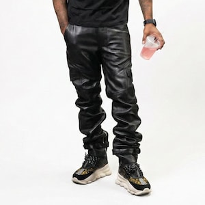 May include: Black leather pants with cargo pockets and a gathered design. The pants are paired with black and gold sneakers. A person is holding a clear plastic cup with a pink beverage.