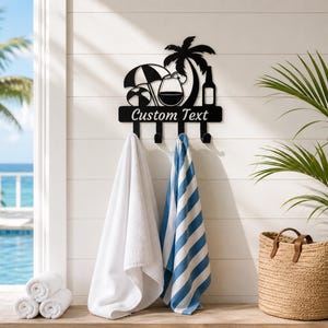 Peut inclure: Un porte-manteau mural en métal noir au design tropical avec un palmier, un parasol, un ballon de plage, un verre à vin et une bouteille. Le support porte l'inscription Custom Text et quatre crochets tenant une serviette blanche et une rayée.