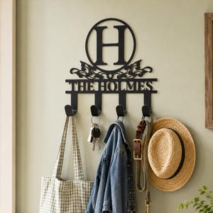 May include: A black metal wall-mounted key and coat rack featuring a circular frame with the letter H, decorative scrollwork, and the text THE HOLMES. The rack includes four hooks holding a checkered tote bag, keys, a denim jacket, and a hat.