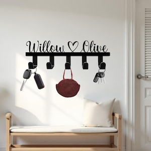 May include: Black metal key holder with the names "Willow" and "Olive" in cursive script, separated by a heart symbol. The key holder has five hooks, with keys and a small purse hanging from them. The key holder is mounted on a white wall.