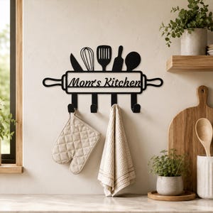 May include: A black metal wall-mounted hook rack shaped like a rolling pin with kitchen utensil silhouettes on top. The center features the text "Mom's Kitchen" in a cursive font. Four hooks hang below, holding a beige quilted oven mitt and a towel.