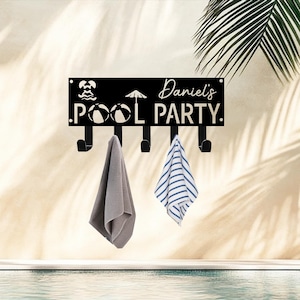 May include: Black metal wall-mounted pool party sign with hooks. The sign has the text "Daniel's POOL PARTY" and pool-themed icons. Two towels hang from the hooks, one gray and one blue and white striped.