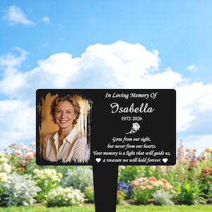 May include: A black memorial plaque with a photo of a woman, the name "Isabella", and dates 1972-2026. The plaque includes the text "In Loving Memory Of" and a memorial quote. It is placed in a garden setting.
