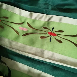 May include: A fabric garment featuring a dark green section and a lighter lime green band. The lime green band is decorated with embroidered floral motifs in brown, pink, and red. The fabric has a smooth, slightly reflective finish throughout.