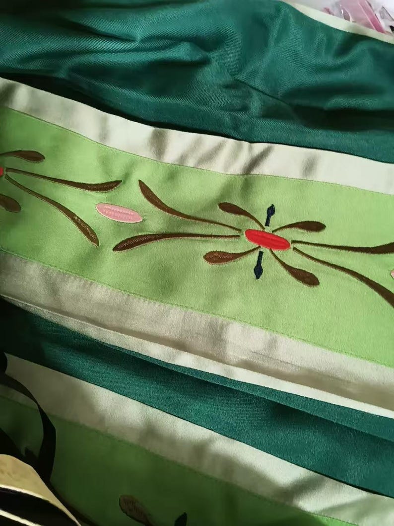 May include: A fabric garment featuring a dark green section and a lighter lime green band. The lime green band is decorated with embroidered floral motifs in brown, pink, and red. The fabric has a smooth, slightly reflective finish throughout.