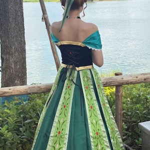 May include: A floor-length dress featuring a black bodice with gold trim and an off-the-shoulder teal neckline. The skirt is dark green with light green vertical panels decorated with a repeating floral pattern in red and pink embroidery.