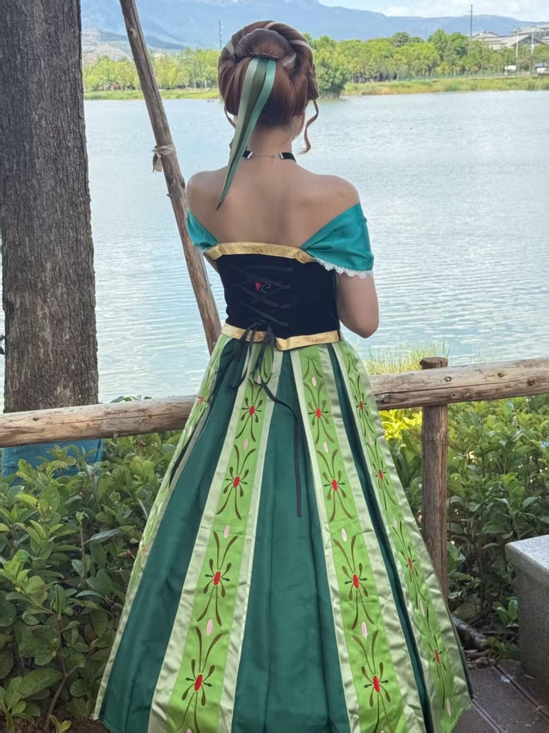 May include: A floor-length dress featuring a black bodice with gold trim and an off-the-shoulder teal neckline. The skirt is dark green with light green vertical panels decorated with a repeating floral pattern in red and pink embroidery.