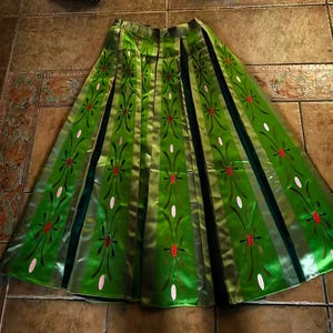 May include: A floor-length green skirt featuring vertical panels with a repeating floral pattern in red and white. The skirt has a flared silhouette with dark green inserts between the panels, creating a structured, pleated appearance throughout.