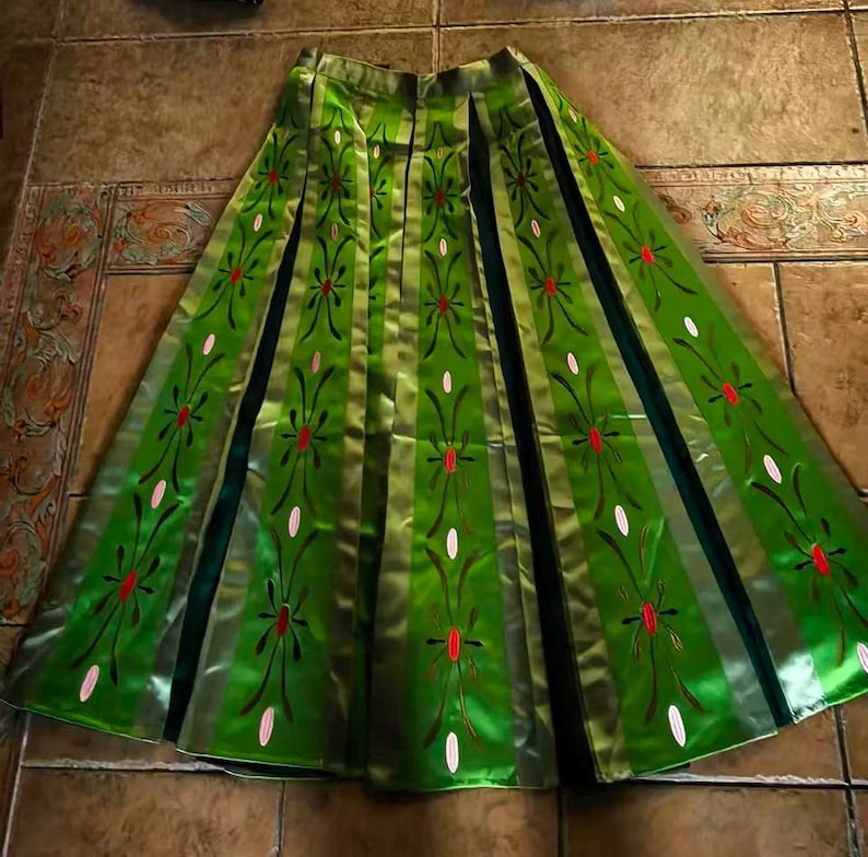 May include: A floor-length green skirt featuring vertical panels with a repeating floral pattern in red and white. The skirt has a flared silhouette with dark green inserts between the panels, creating a structured, pleated appearance throughout.