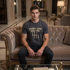 May include: A dark grey t-shirt with the text "MANUAL GEAR REAL FEEL" and a gear shift pattern in gold. The shirt is worn by a person sitting on a beige sofa. The person is wearing dark jeans.