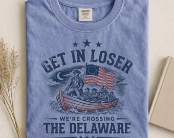 Può includere: T-shirt blu con la scritta "GET IN LOSER" sopra un'illustrazione di una barca che attraversa un fiume, con il testo "WE'RE CROSSING THE DELAWARE" sotto. Il disegno include una bandiera e delle stelle. La maglietta ha un'etichetta Comfort Colors.